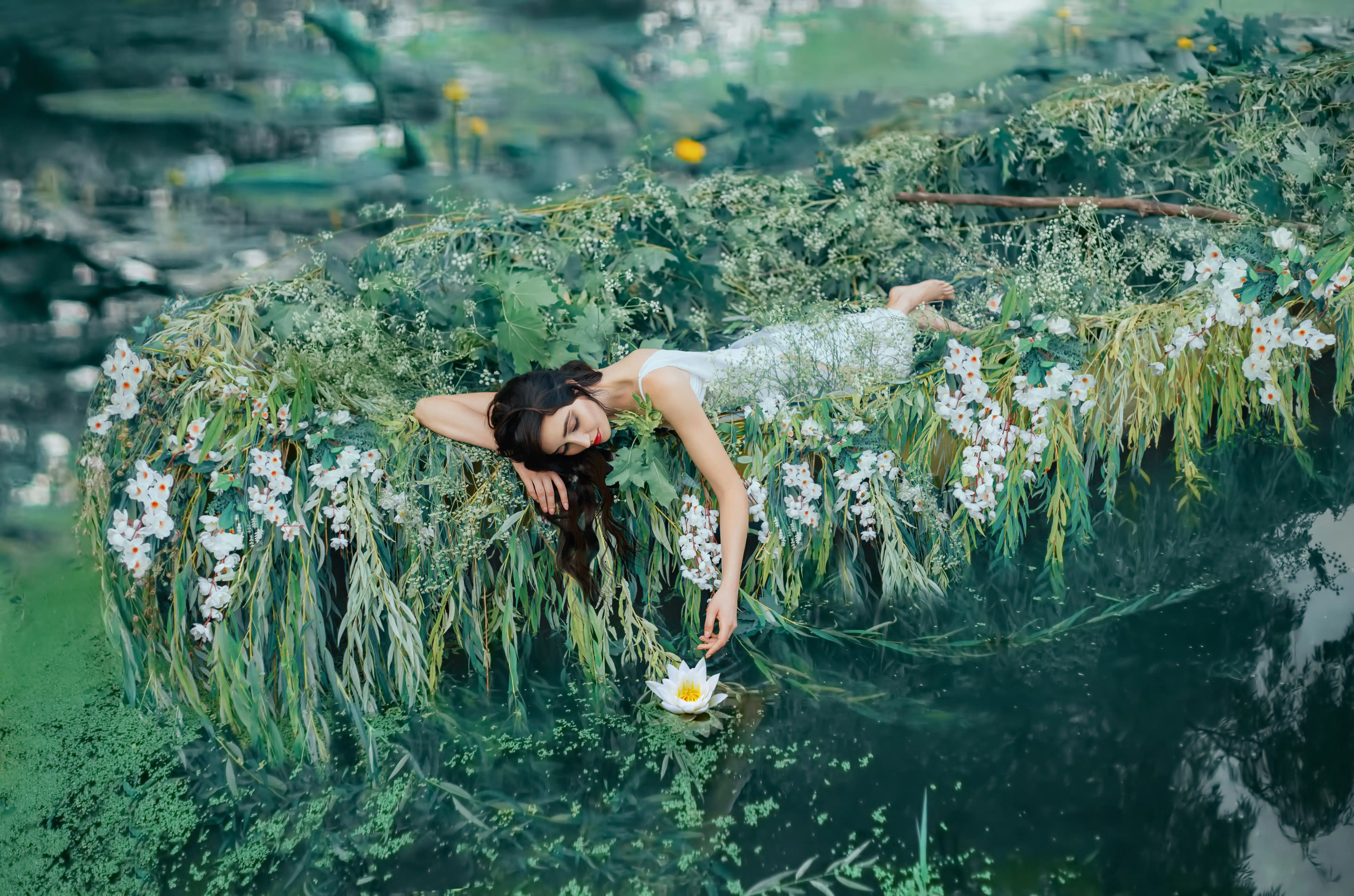 A brunette woman with long hair lies in a boat and gently touches a water lily her hand.