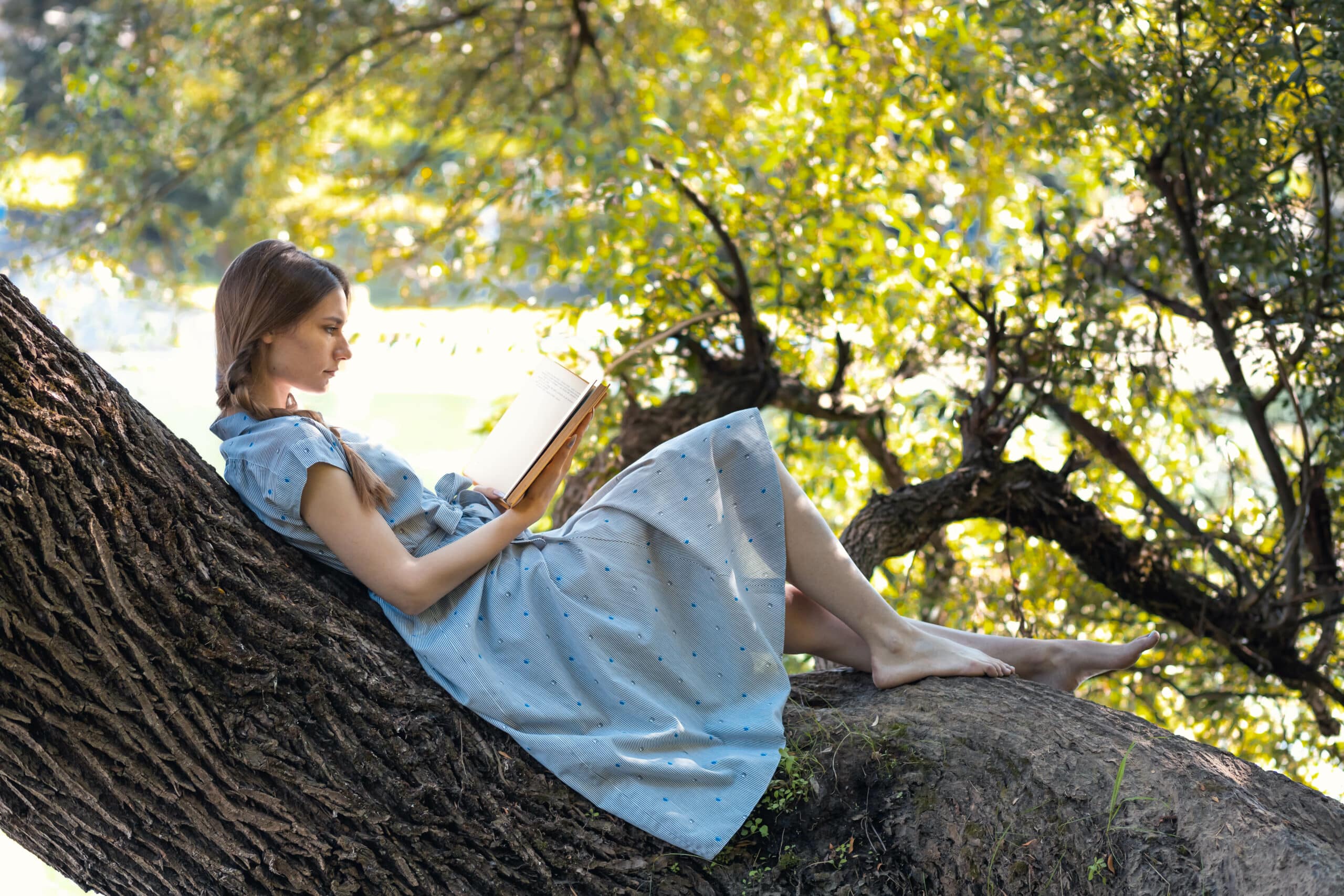 Young woman sitting on a tree by the river, reading a book.