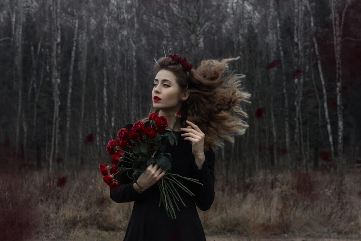 A beautiful girl with curly blond hair stands in a black dress with flowers.