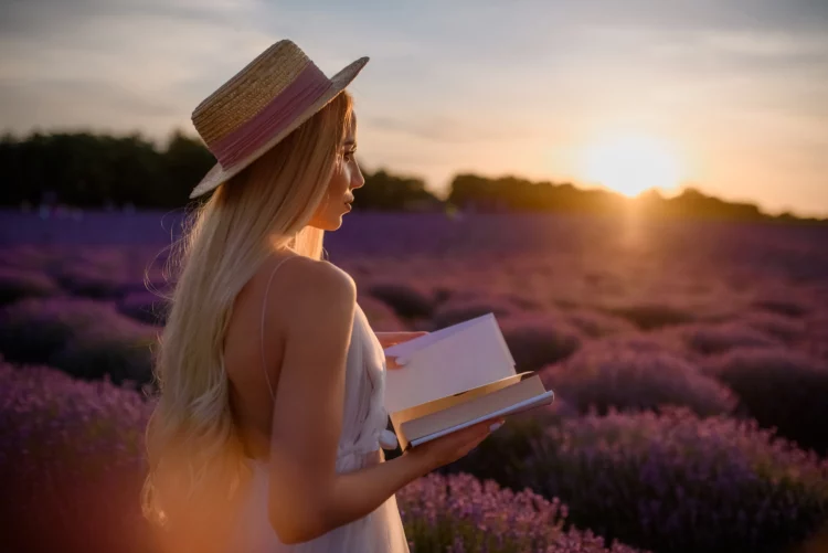 Young beautiful woman in white dress and hat is walking in a lavender field with a book