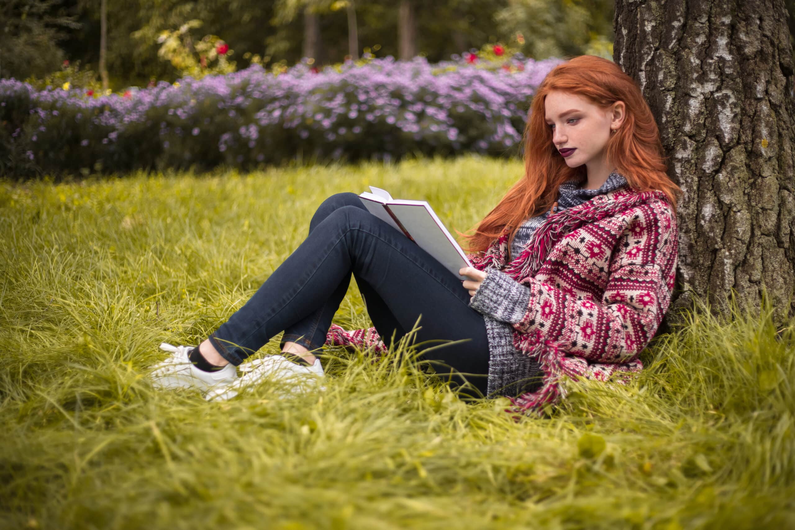 young redhead woman with freckles and dark lipstick reading book outdoors in park under the big tree on grass with flowers.