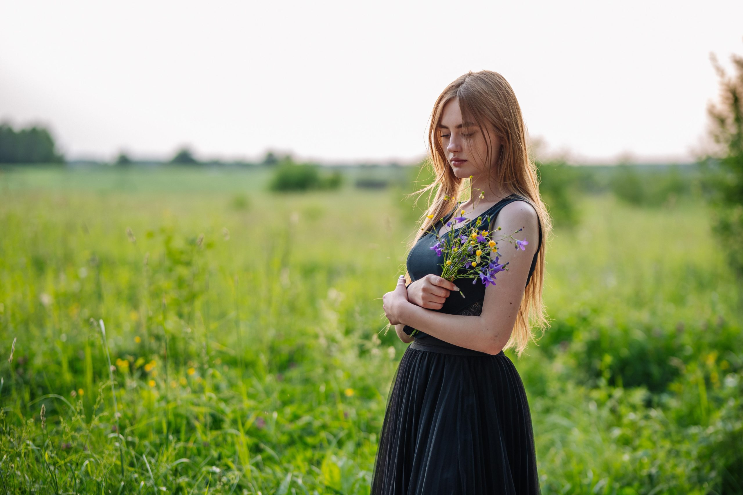 Attractive sad-looking girl hugging a bouquet of wild flowers at sunrise outdoors.