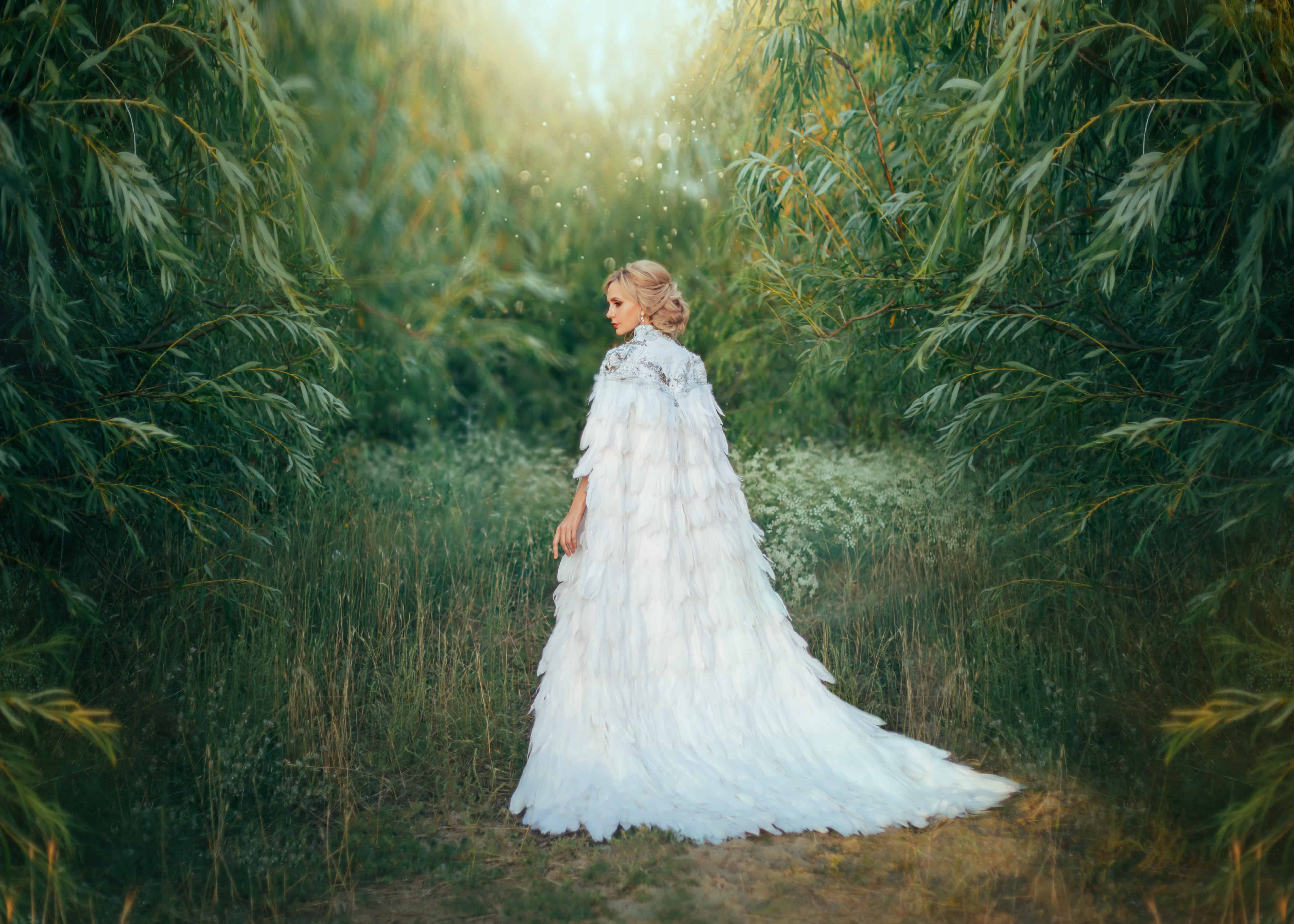 Fantasy woman Queen in white medieval dress with feathers. Creative clothes long cape, royal, vintage cloak with train. Greek Goddess swan bird. Art photography. Green forest tree. image trendy bride
