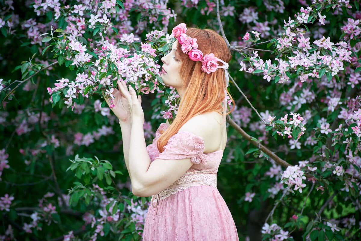 A young woman in a pink lace dress and a wreath of roses smells the flowers in the garden