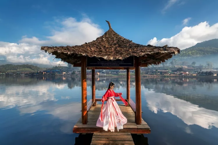Woman wearing chinese traditional dress standing on wooden foot bridge in the middle of a picturesque lake.