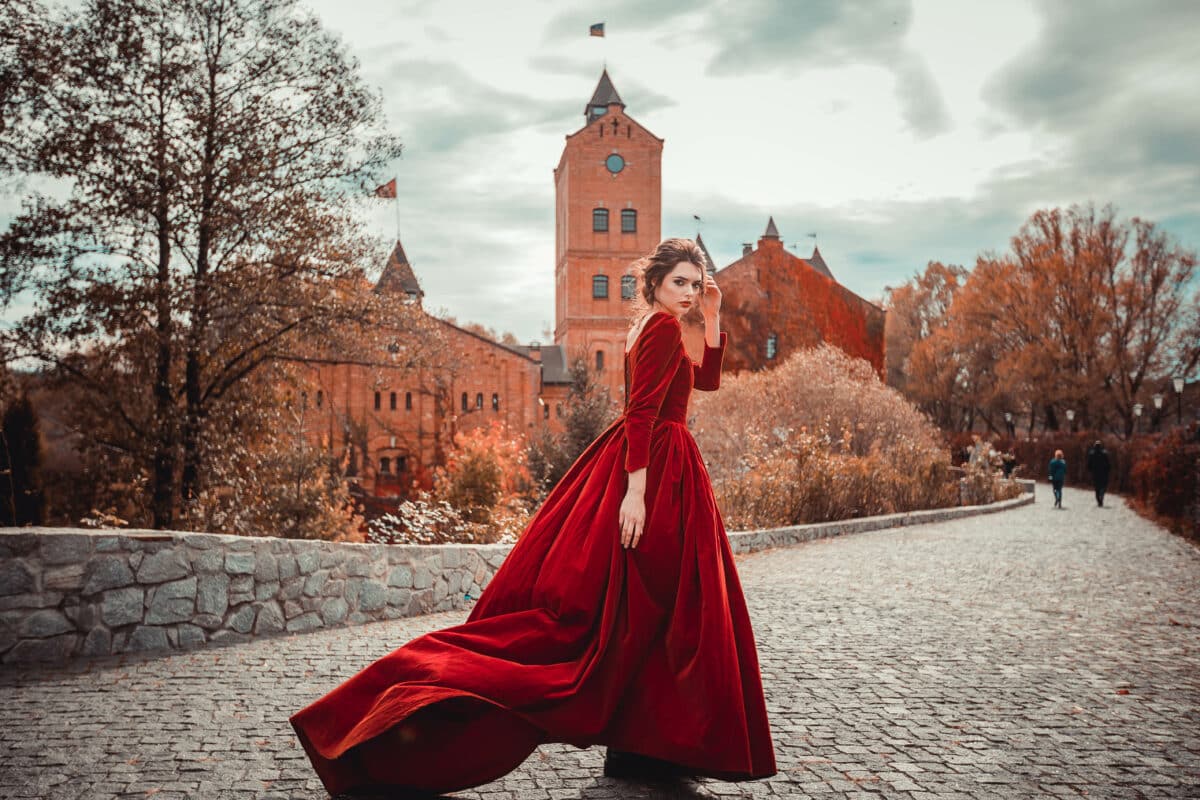 Beautiful girl in a burgundy red dress walking near old castle on a background of autumn grape leaves in the park,