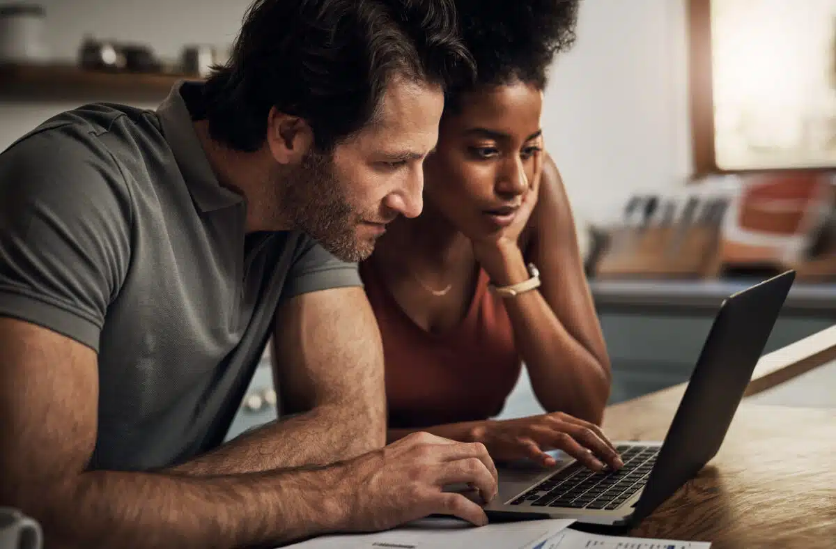 Couple, finance and laptop in a kitchen for planning, budget and savings or paying bills together in their home. Interracial marriage, online and people with tax, mortgage or home loan or insurance.