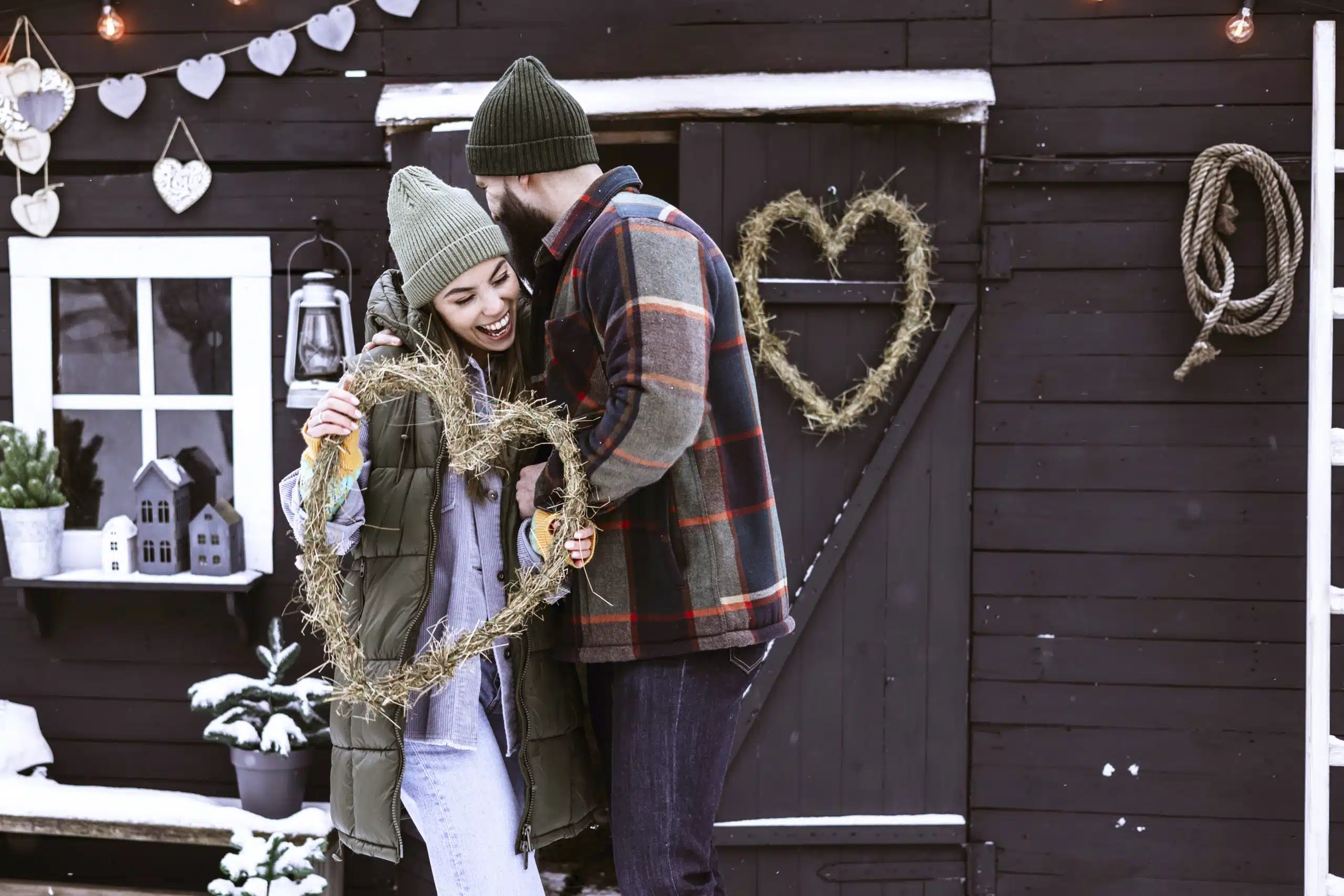 Young couple in love in stylish winter clothes standing near porch of country house with handmade wreath of dried grass in shape heart.