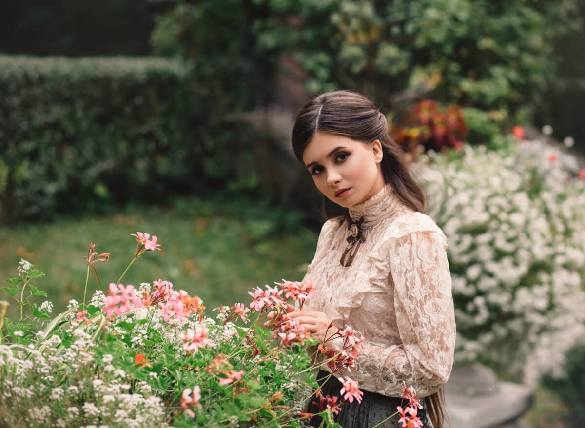 A girl walks in a flowering garden, she has a vintage blouse with a bow, chestnut long hair. she gently cares for her flowers. Sweetheart gardener. Artistic photography. Love of plants