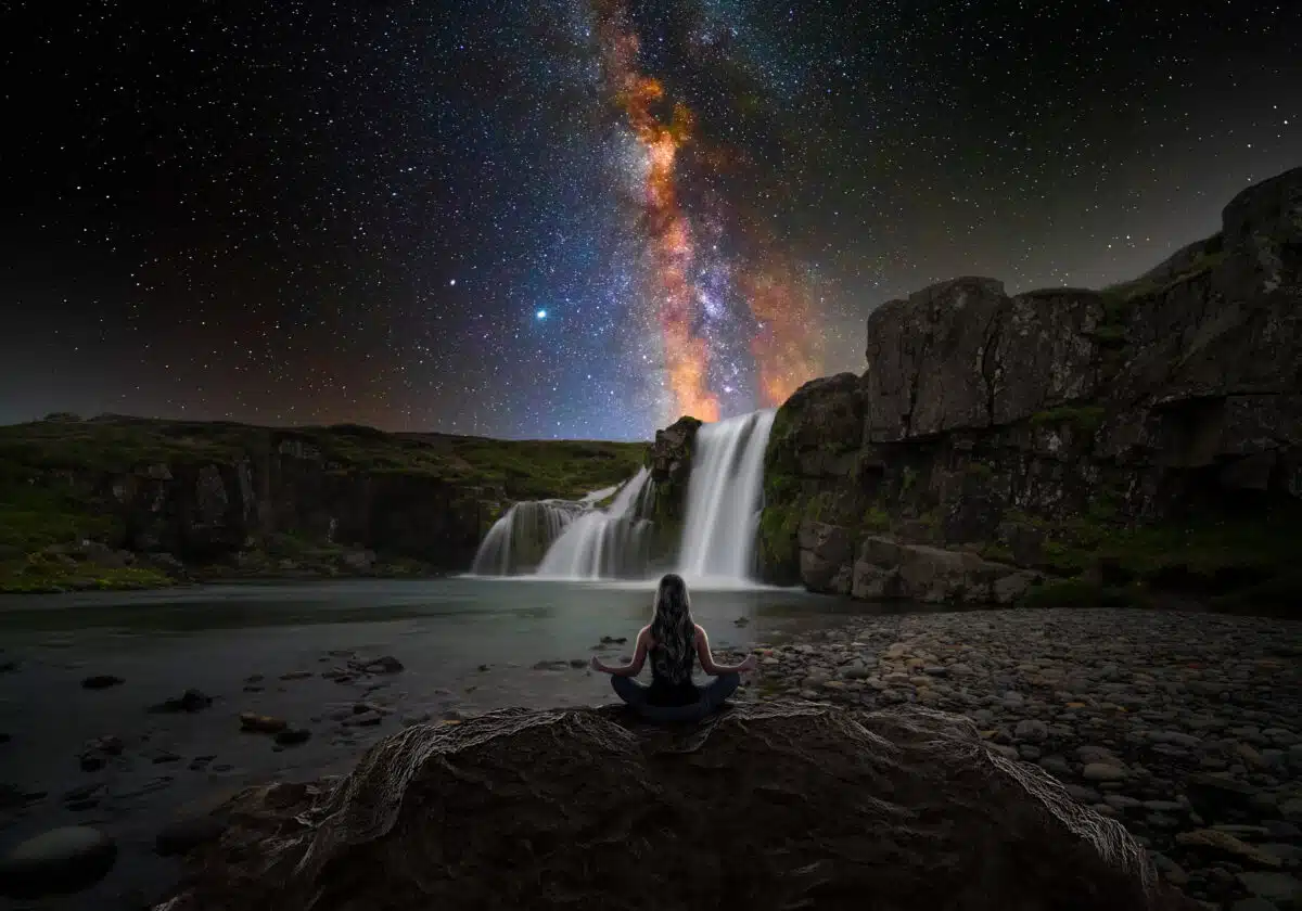 a woman sitting in lotus position in front of a magic waterfall and the milky way stars