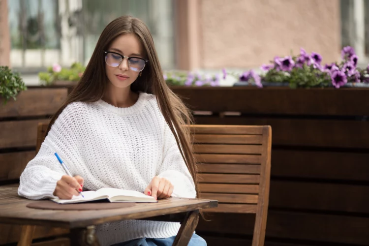 urban female writer sitting in a cafe wearing glasses writing in her notepad.