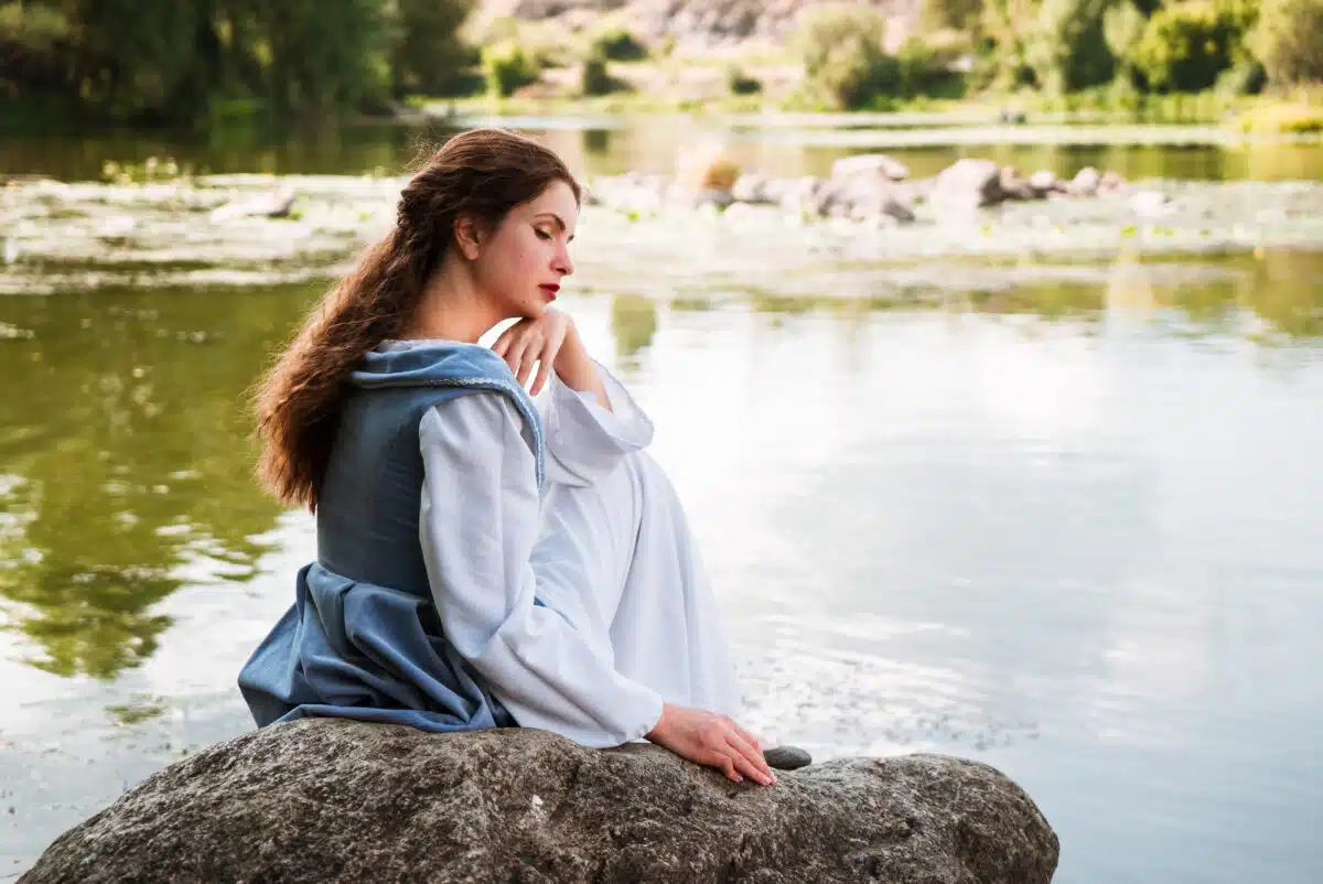 a woman with long hair in a traditional blue dress sits by the river