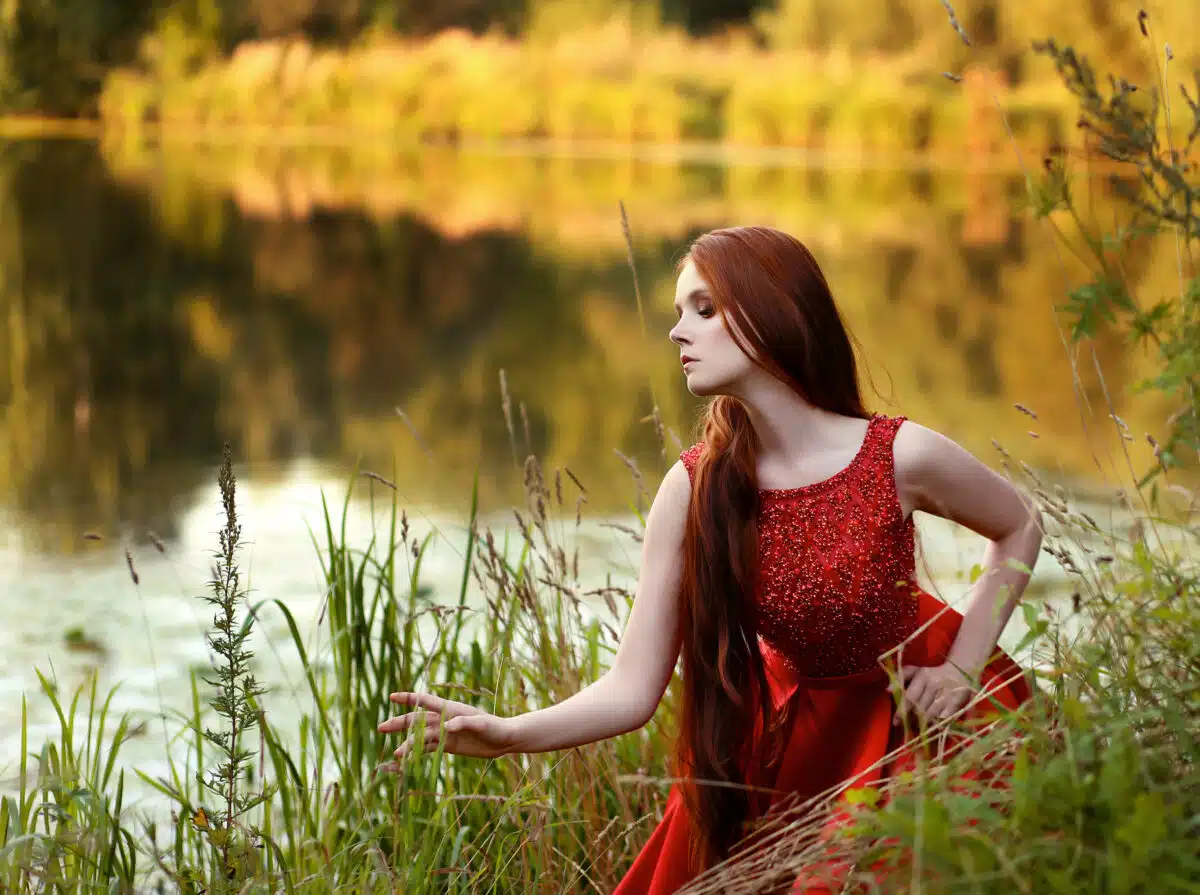 Beautiful red head lady in red evening dress near lake in autumn