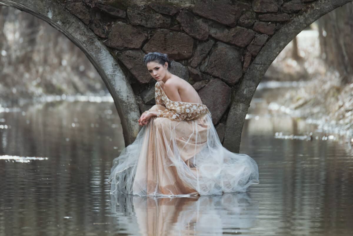 Atmospheric outdoor portrait of sensual young woman wearing elegant dress sitting under the bridge of river.