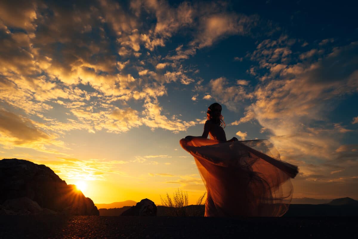 a woman in a fluttering dress stands on a mountain against the backdrop of a sunset sky