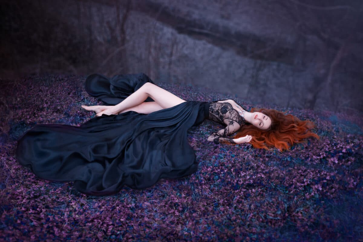 lady sleeping on the ground with long red fiery hair lies in a luxurious black lace dress with a magnificent train. poisoned princess with sexy bare legs fell on purple forest flowers, creative photo
