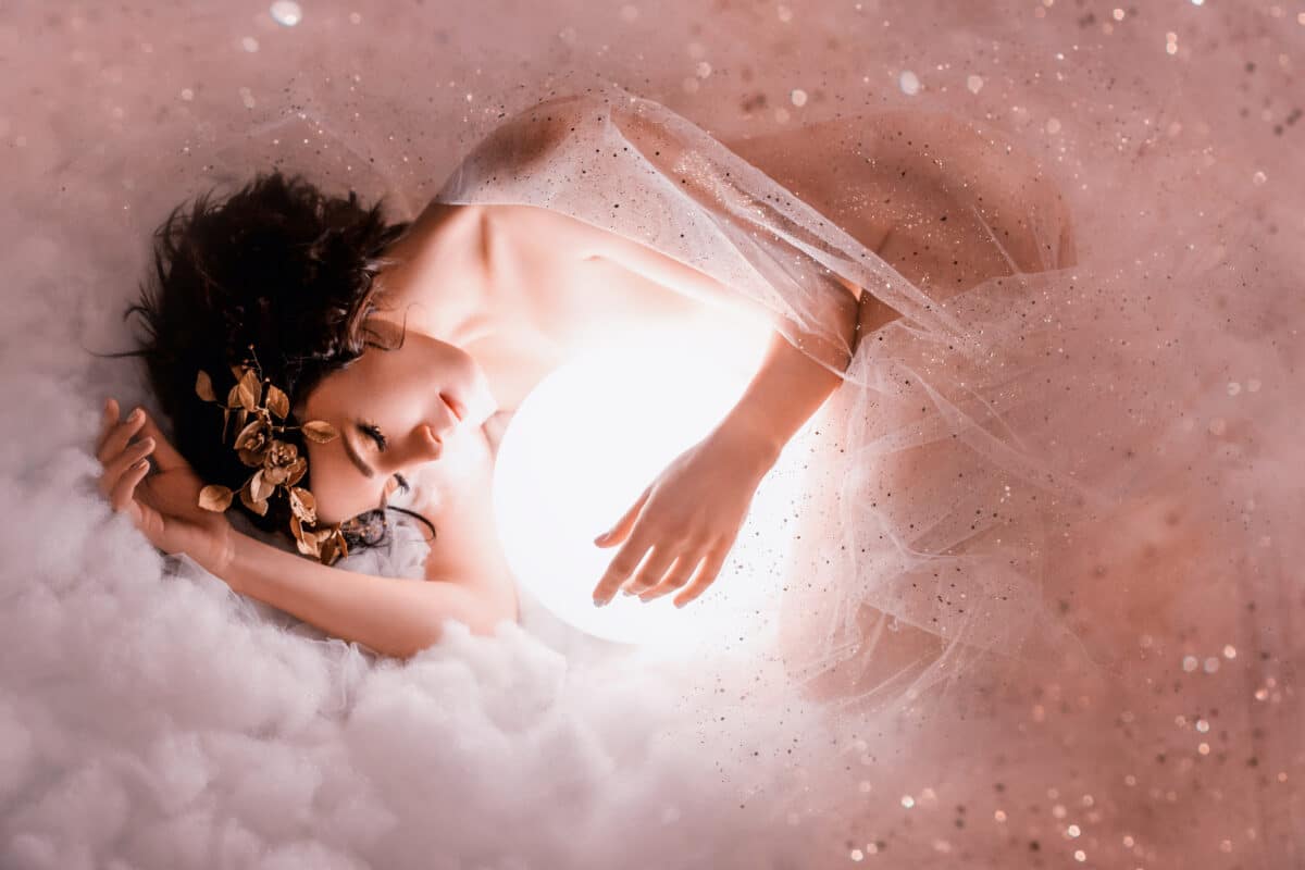 gentle image of angel sleeping next to the moon in pink light clouds, naked body of slim girl lying in a mist, covered with glitter sequin, princess of the night with dark hair and a wonderful wreath