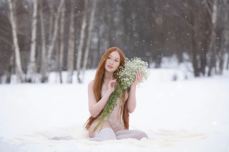 Red-haired woman sitting on the snow with a bouquet of white flowers.