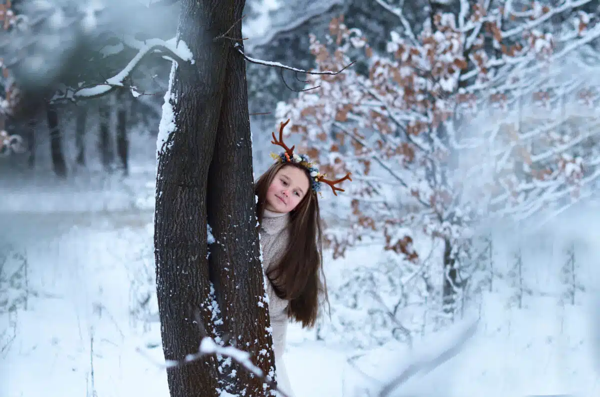 fairy girl with deer horns hiding behind a huge tree trunk in the winter forest