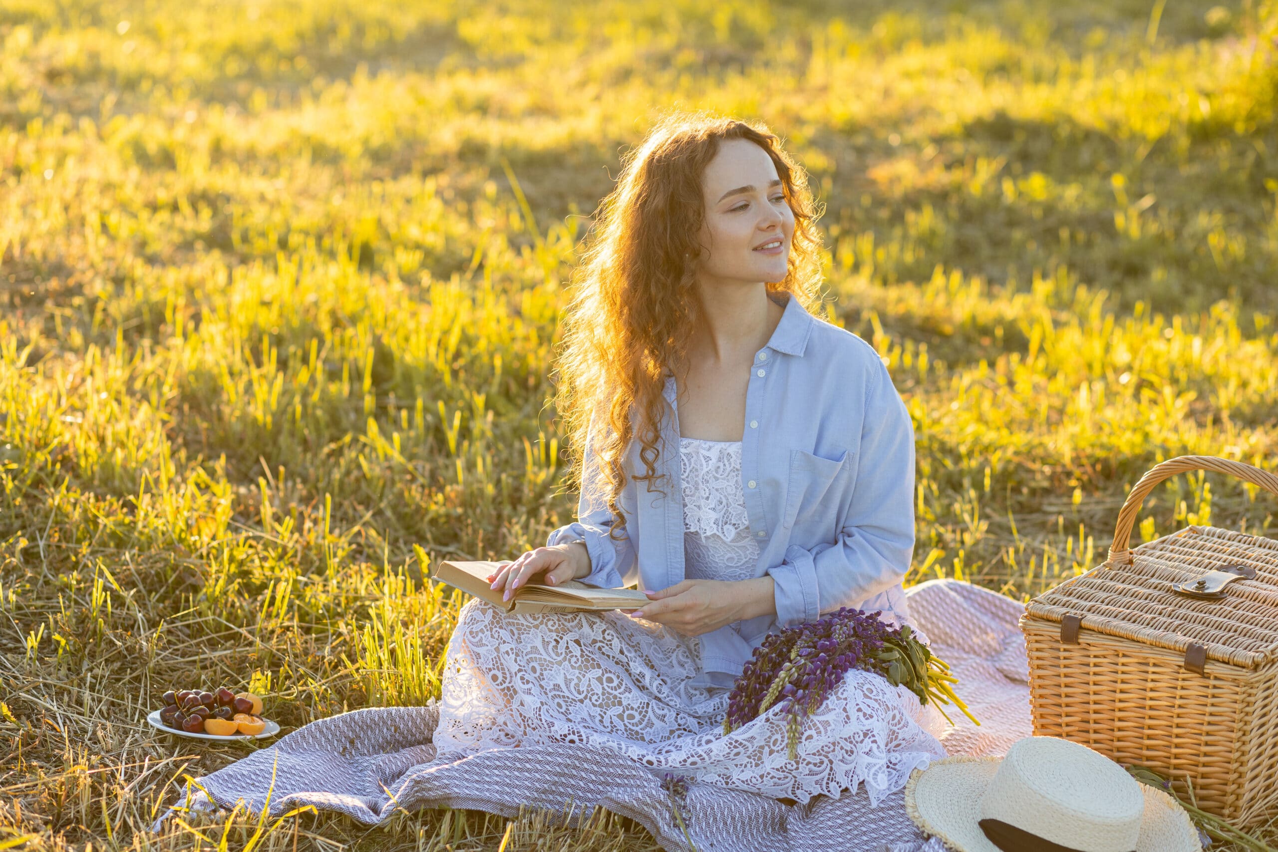 Beautiful young girl in a white dress, straw hat, picnic basket reading a book on a meadow.