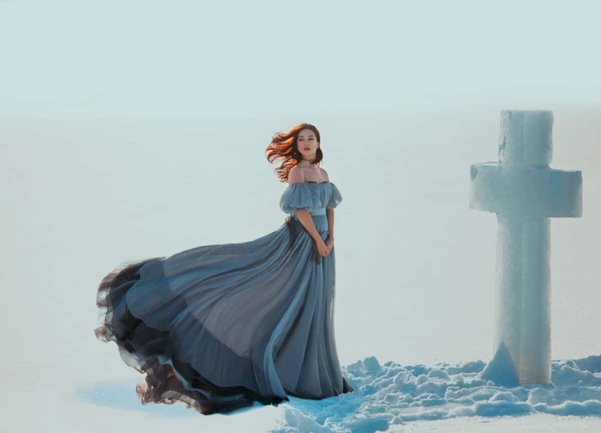 Young sad woman princess. Backdrop blue sky white snow winter. Snowy frosty desert Ice cross. Religion Concept spirituality pray with hope. Red hair flying in wind. Medieval vintage dress fluttering