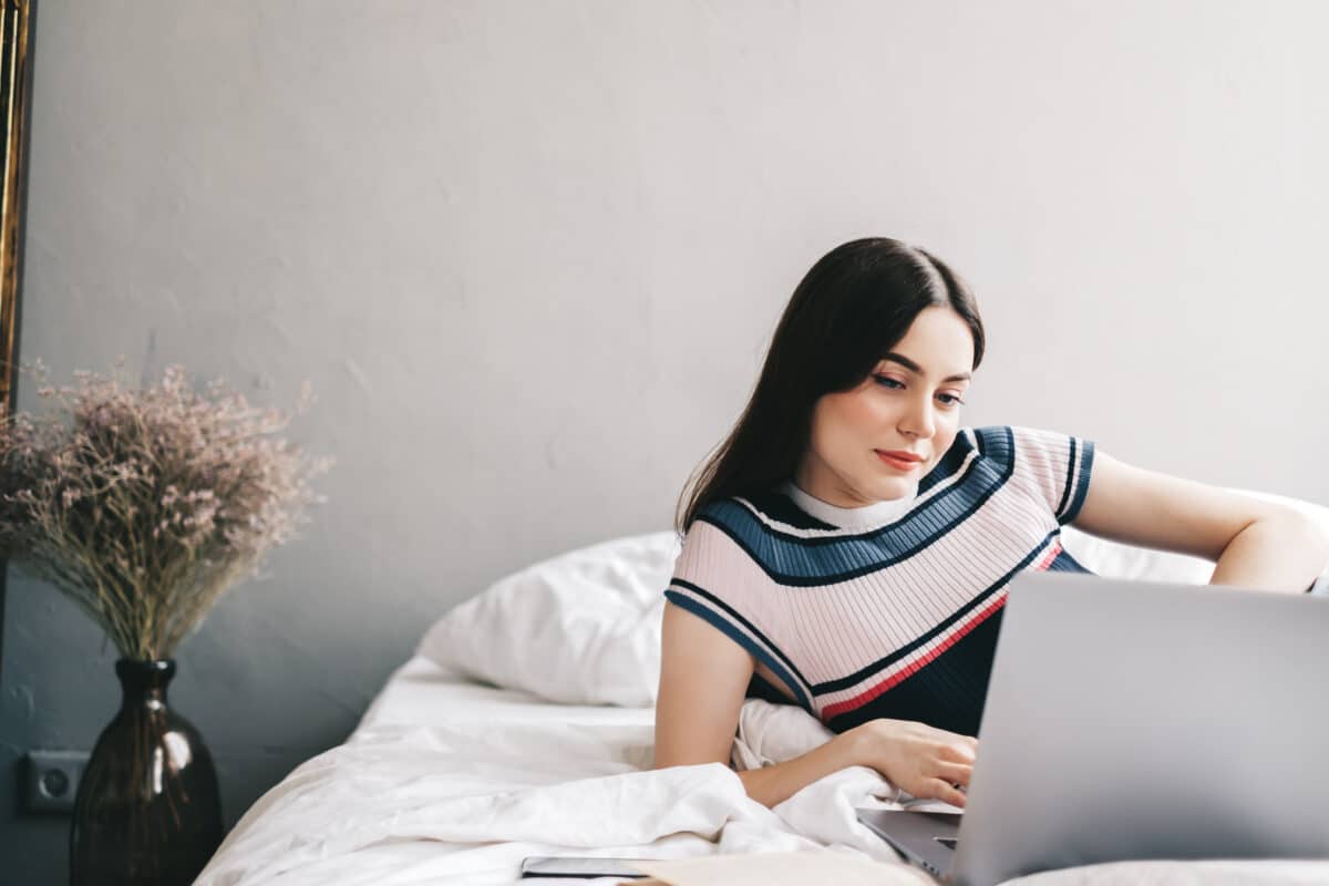 Beautiful young caucasian woman resting on a bed at home and using laptop computer. Chatting with friends, shopping online, distance education.