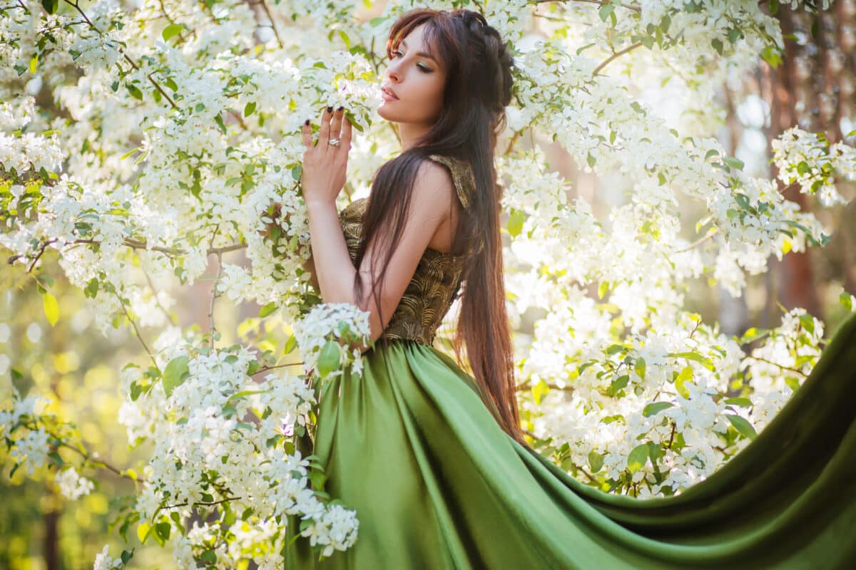A beautiful young woman in a green dress with a large train among the blooming apple trees.