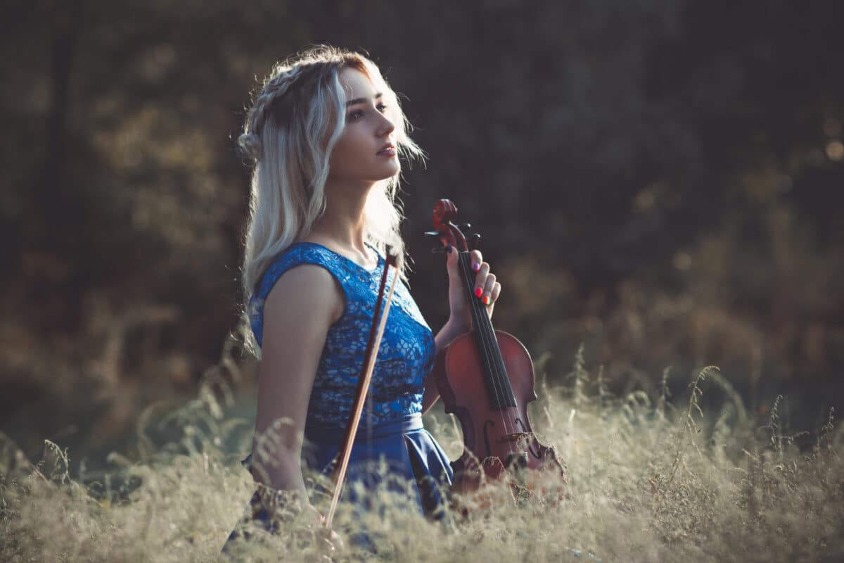 portrait of a young fabulously beautiful girl in a dress with a violin sitting in dry grass on meadoe at the dawn, woman playing a musical instrument with inspiration relaxing on nature