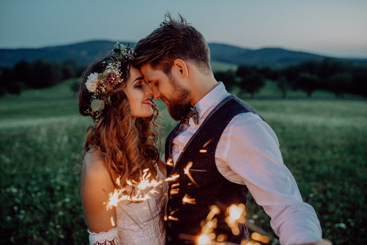 Beautiful young bride and groom on a meadow in the evening, holding sparklers.
