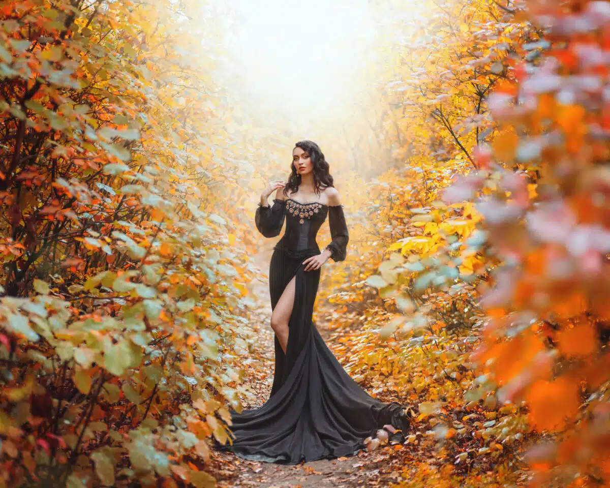 Dark Queen woman stands in mystical autumn forest. Orange foliage gothic trees mis. Art Fantasy fairy tale princess girl. Vintage long royal sexy black golden dress, puffy sleeves open bare