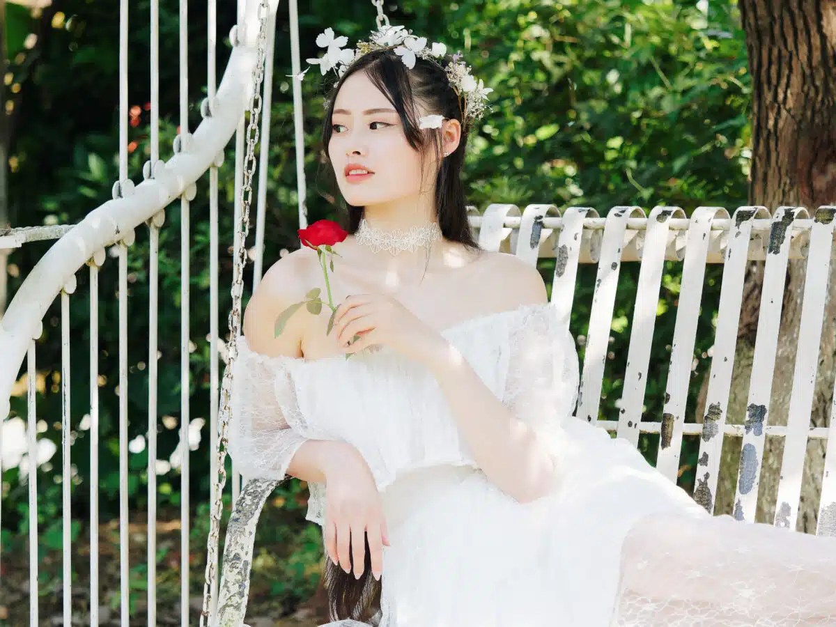Beautiful fairy lady in white wedding dress and garlands of flowers, posing with red rose in hand, pretty Chinese girl sit in white chair in sunny day. Emotions, people, beauty and lifestyle concept.