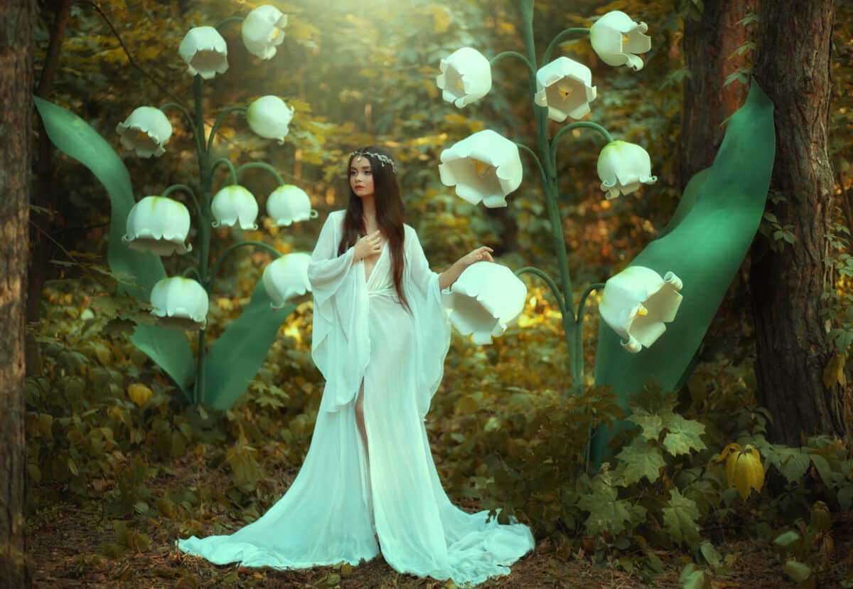Beautiful fantasy woman princess elf in long white dress walks in fairy forest with large flowers lilies of valley. Queen girl in silver diadem. Silk vintage outfit with wide sleeves. Green trees