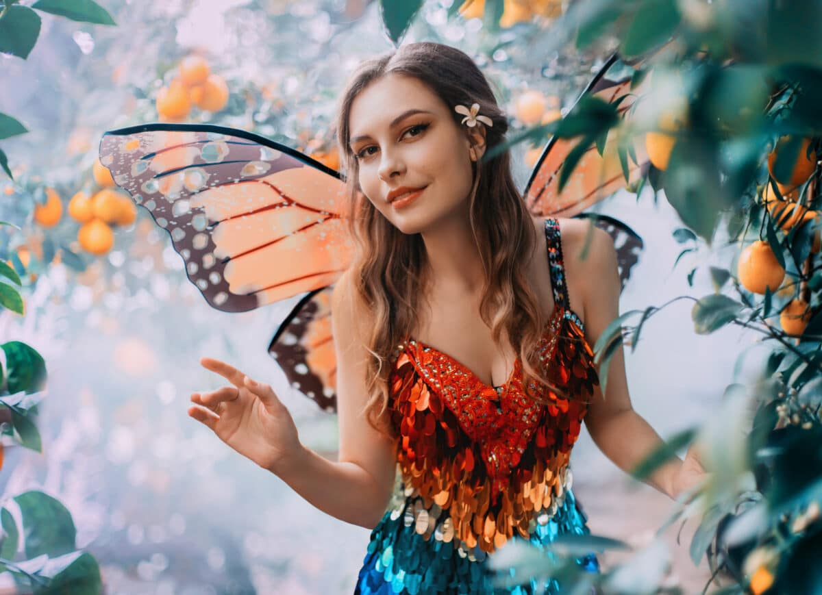 fantasy happy woman fairy walks in jungle. Happy girl in carnival costume bright orange monarch butterfly wings. Red shiny dress. Background Garden lemons fruits green tree mystical fog. Smiling face