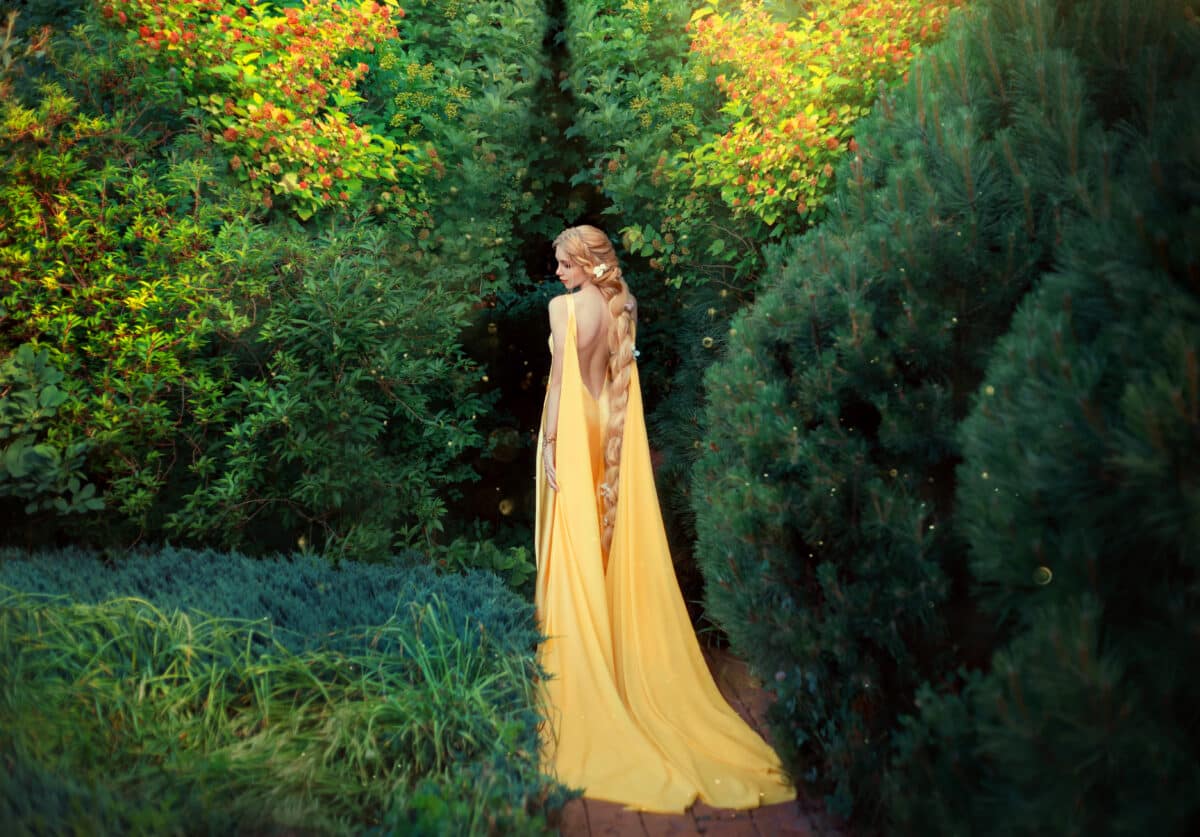 slender beauty in elegant bright dress with stretching trains goes to thick of magical garden, golden elf princess with bare trimmed seductive back and long light braided hair looks at her plants.