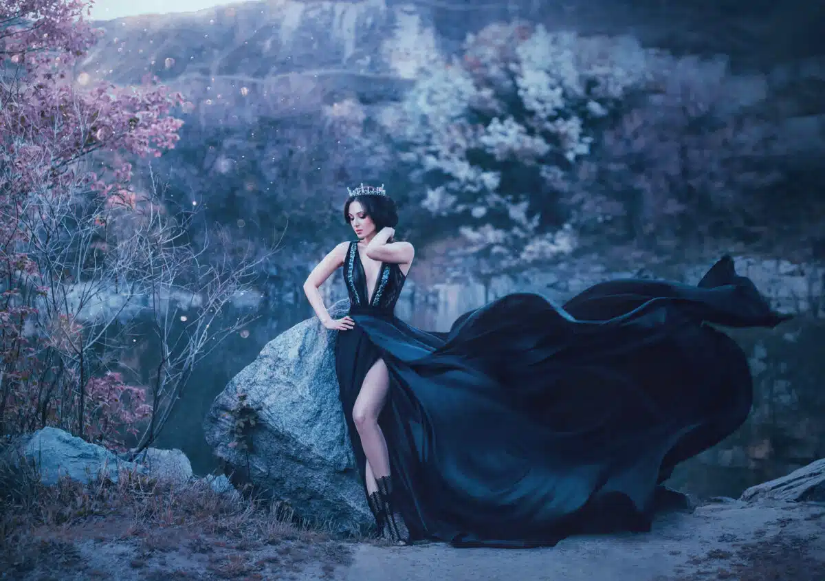 The dark queen pose against the background of gloomy rocks. A luxurious black dress with a long train fluttering in the wind denuding her leg. Elegant, collected hairstyle with a gothic crown.