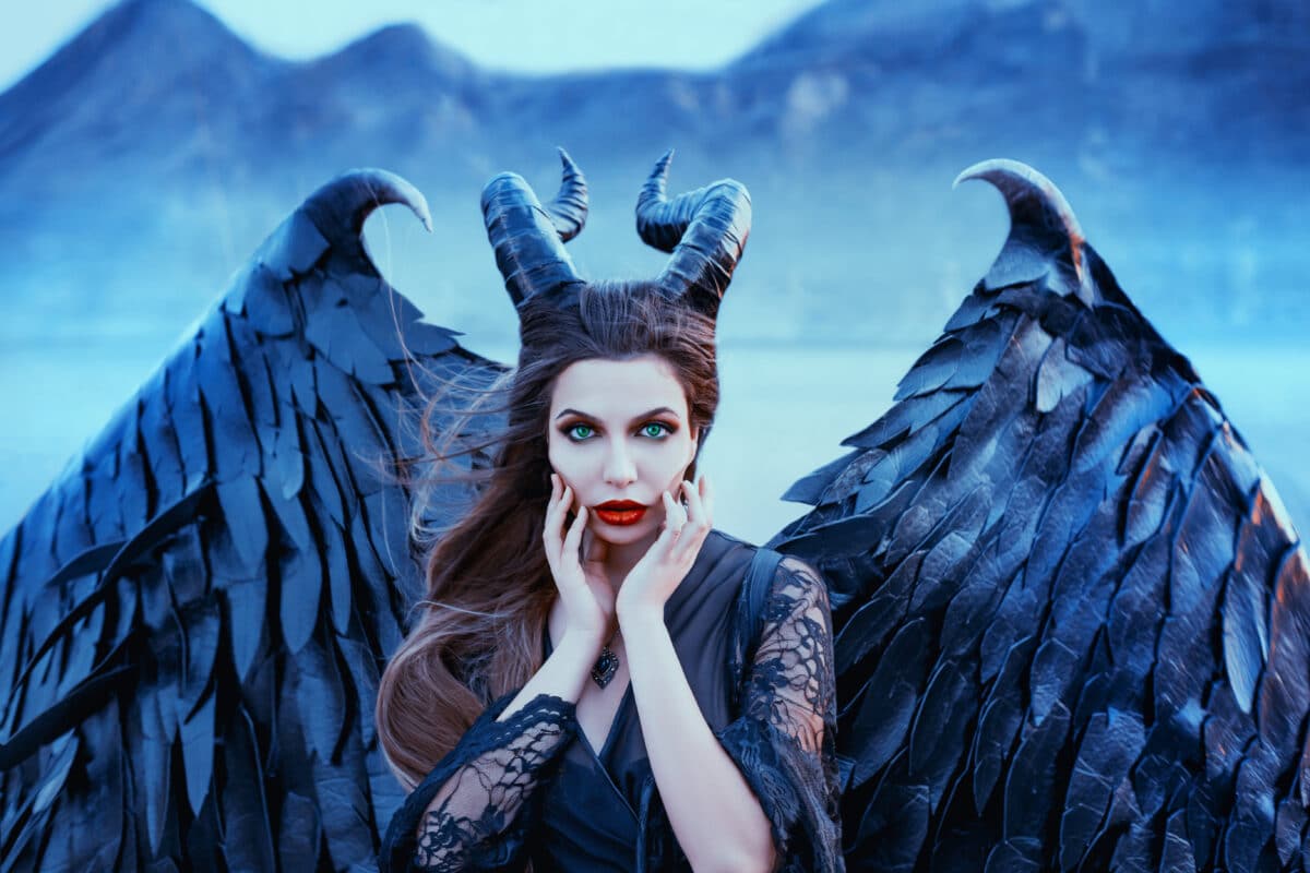 charming portrait of dark angel with sharp horns and claws on strong powerful wings, wicked witch in black lace dress brought hands to face, bright red lipstick and cold eyes, art photo in blue shade