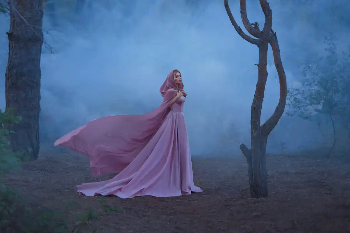 wonderful herbalist enchantress with blond hair, dressed in an expensive luxurious long soft pink dress, holding a bright hood with a hood, the girl stands alone in a dark forest full of fog