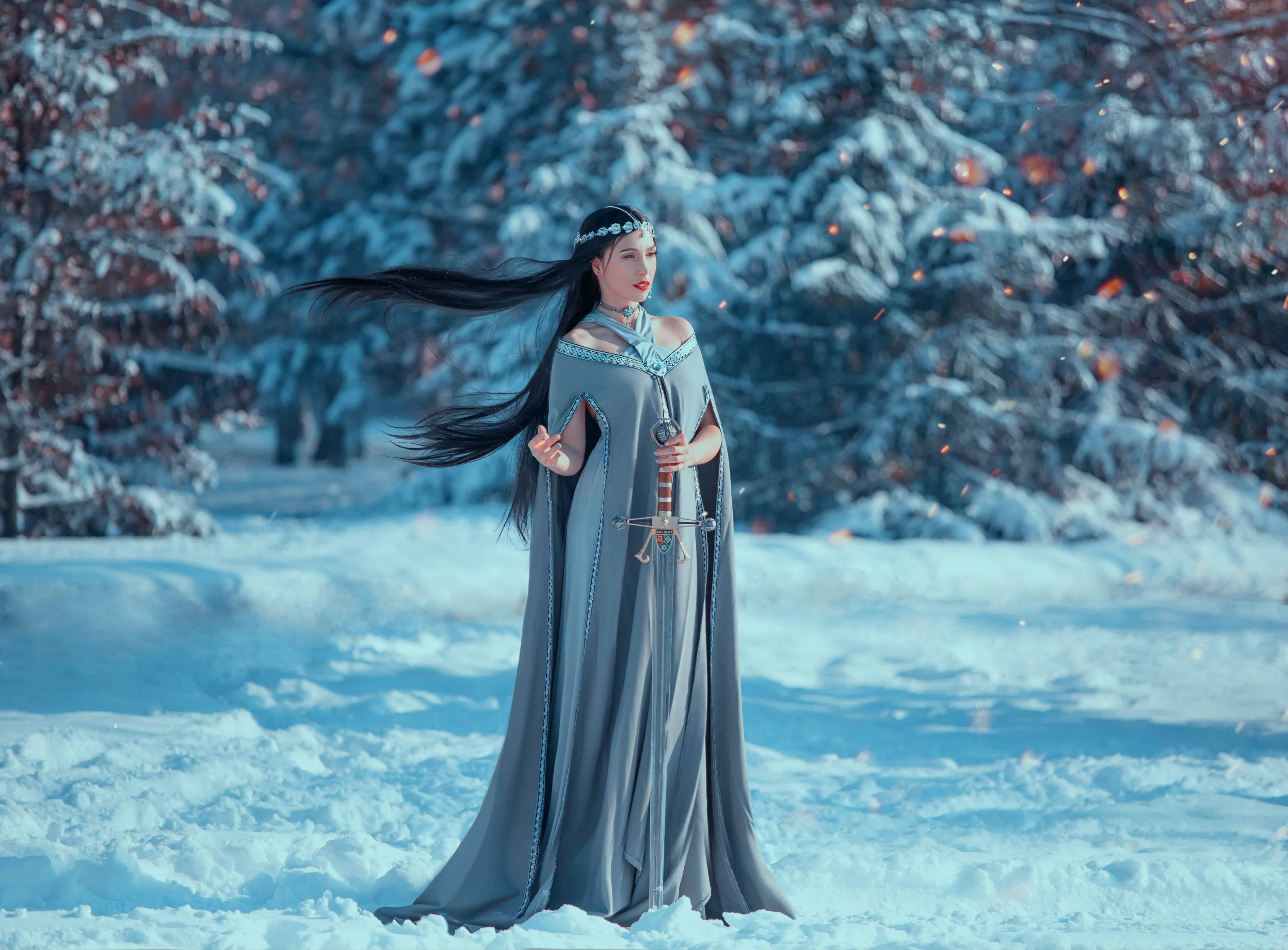 charming attractive lady in snowy forest, militant elf princess with black long flying hair holds sword, loose gray warm dress and raincoat in sparks of fire in winter, creative cold blue colors
