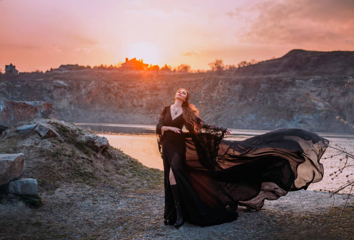 Fantasy woman in black fluttering silk dress standing by the lake at sunset