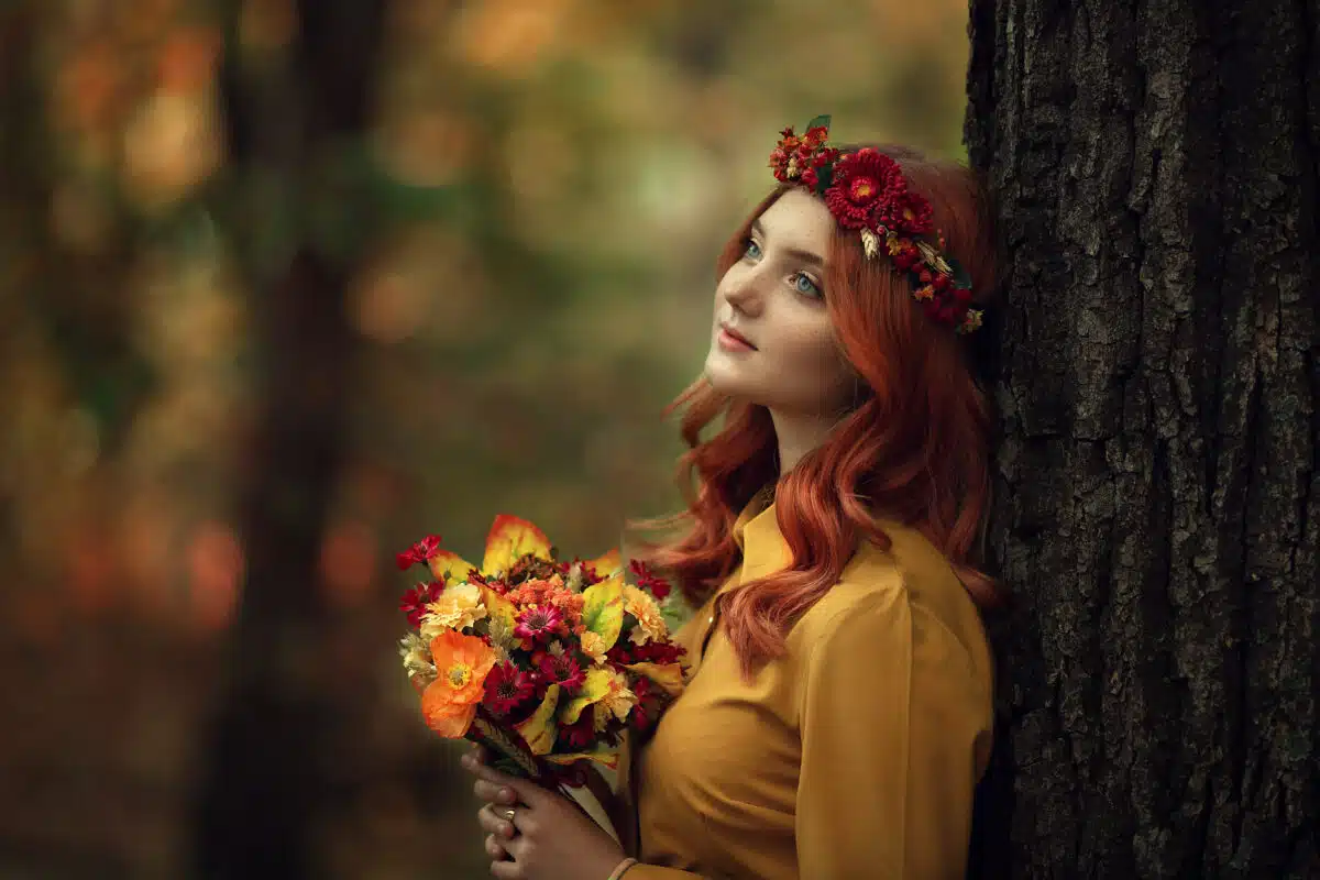 Redhead lady in the autumn forest with flower wreath and bouquet of flowers in her hand
