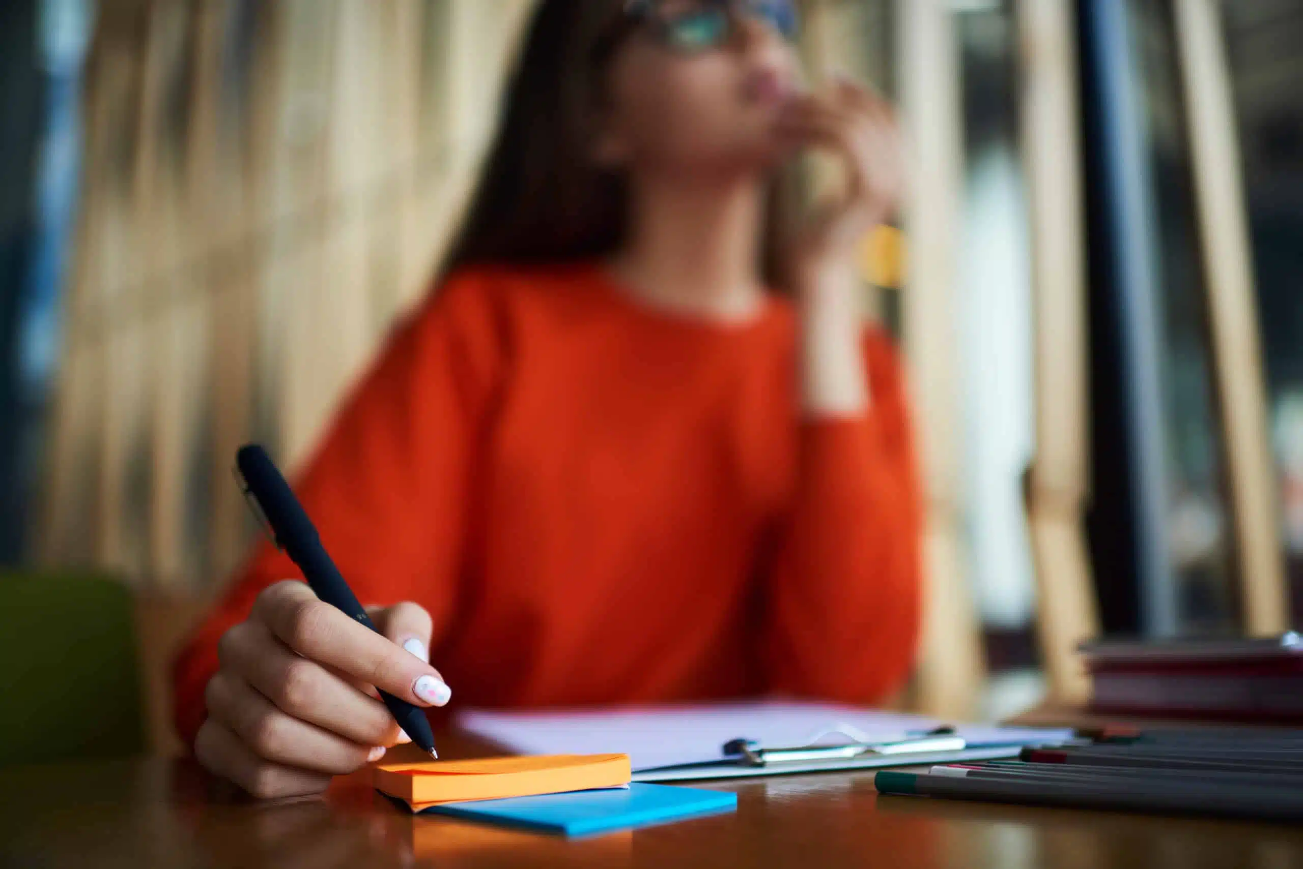 Pensive woman with glasses in red sweatshirt holding a pen, hand resting on her notes.