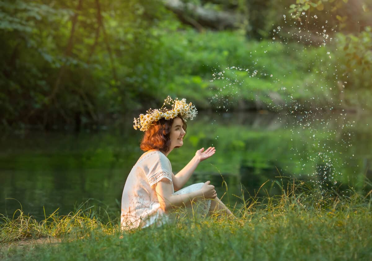 a young beautiful girl in a white dress with hair wreath is sitting by the river in the forest