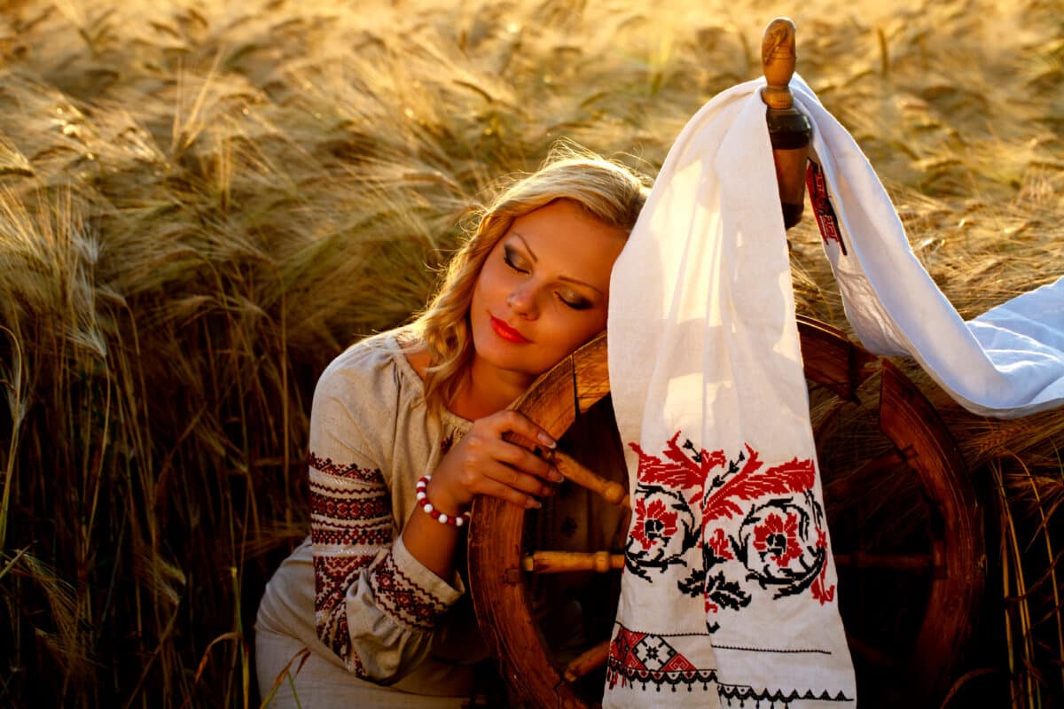beautiful young woman in a traditional dress is sitting in the field with a spinning wheel