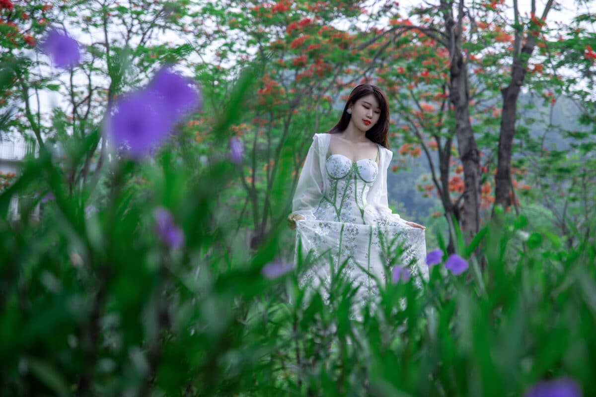 a lady in white long dress walks in the garden with purple blooms