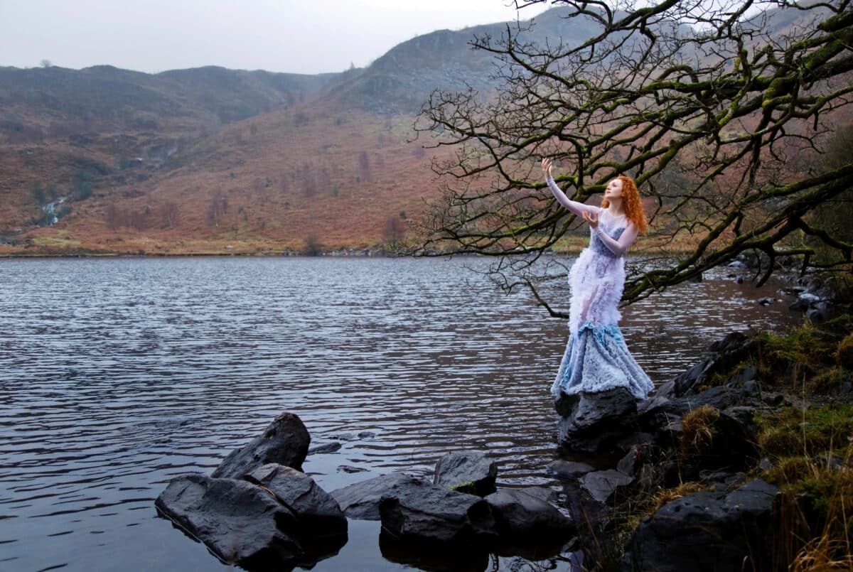 Beautiful red-haired lady dressed in blue white standing on a rock by the lake raising her hands towards the sky as if in prayer