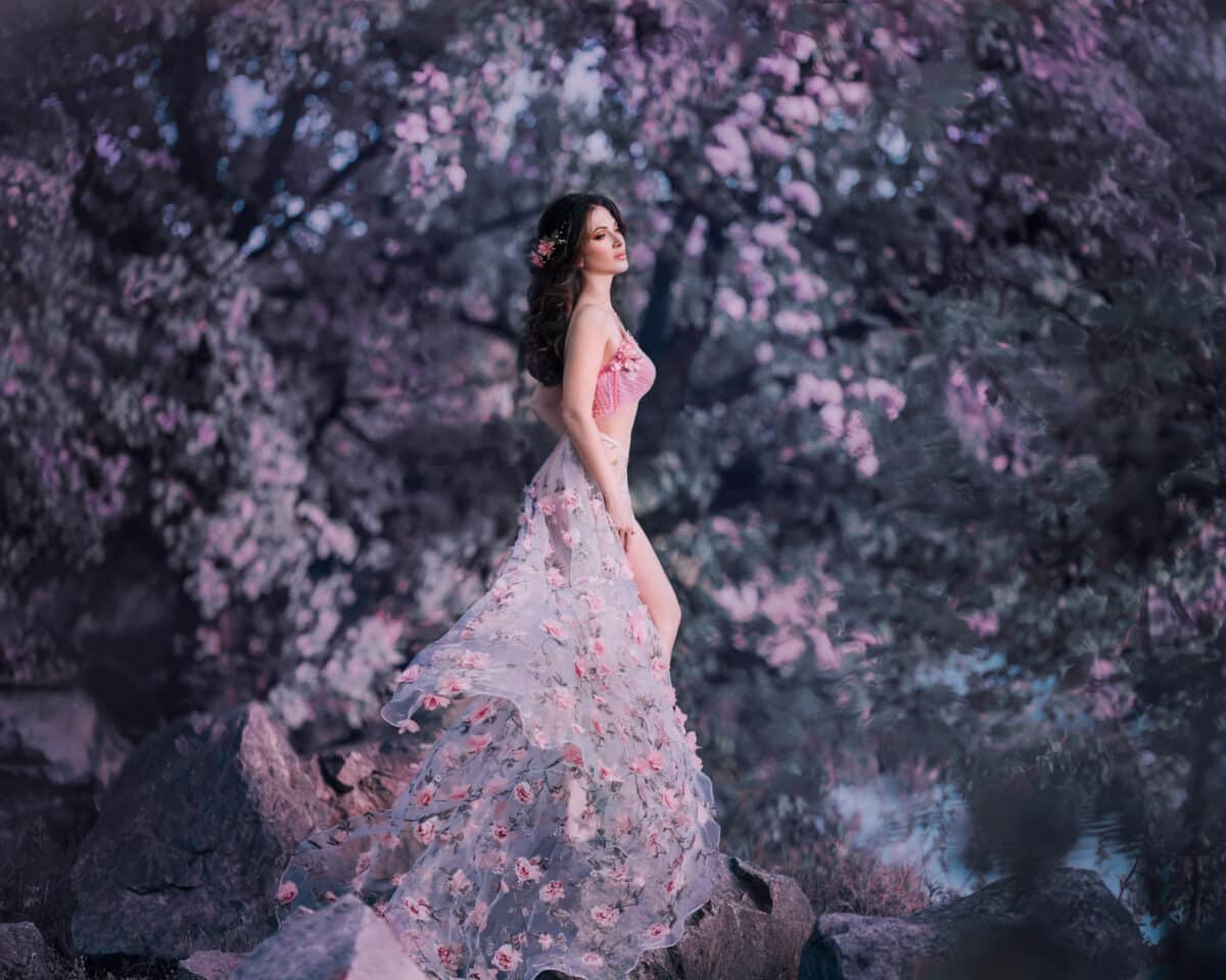 The spring fairy stands on the background of a flowering, rosy tree. She wears a pink dress with flowers that flutters in the wind, emphasizing a sexy silhouette and long legs. Art photo