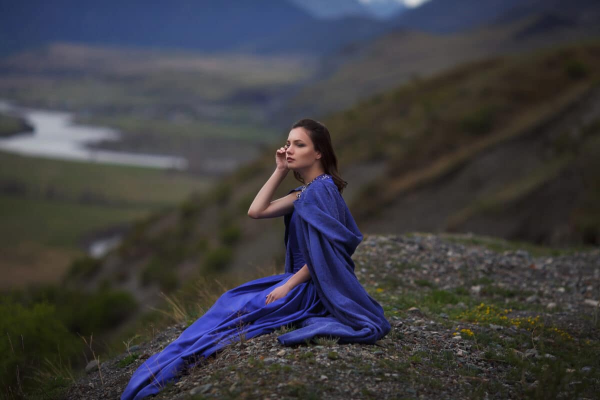 A girl in a magnificent blue dress sits on the edge of a mountain cliff