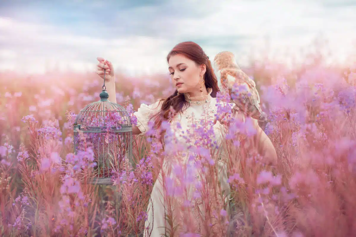 Woman with owl on her hand with cage in other one stay at field of pink flowers.