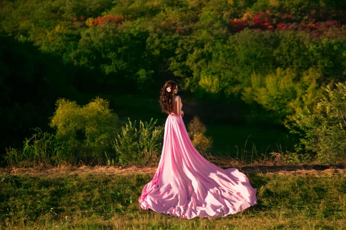The girl in transparent pink dress with a long train flying, decorated with a flower in her hair. Little pink fairy stands on top of the green hills, creative computer color fashionable toning.