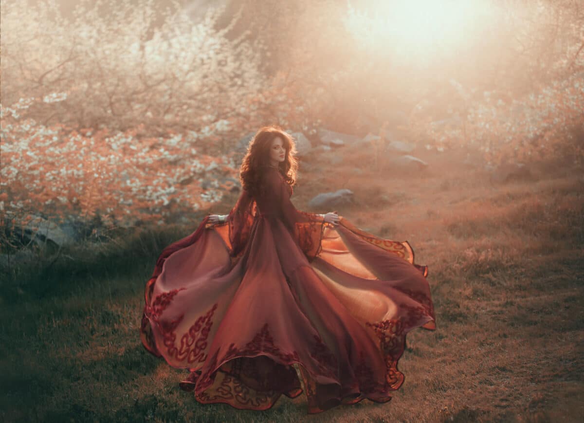 A brunette girl with wavy, thick hair runs to the sun and looks back. The princess has a luxurious, chiffon, red dress that flutters in the wind. Background of a fiery sunset and wild nature